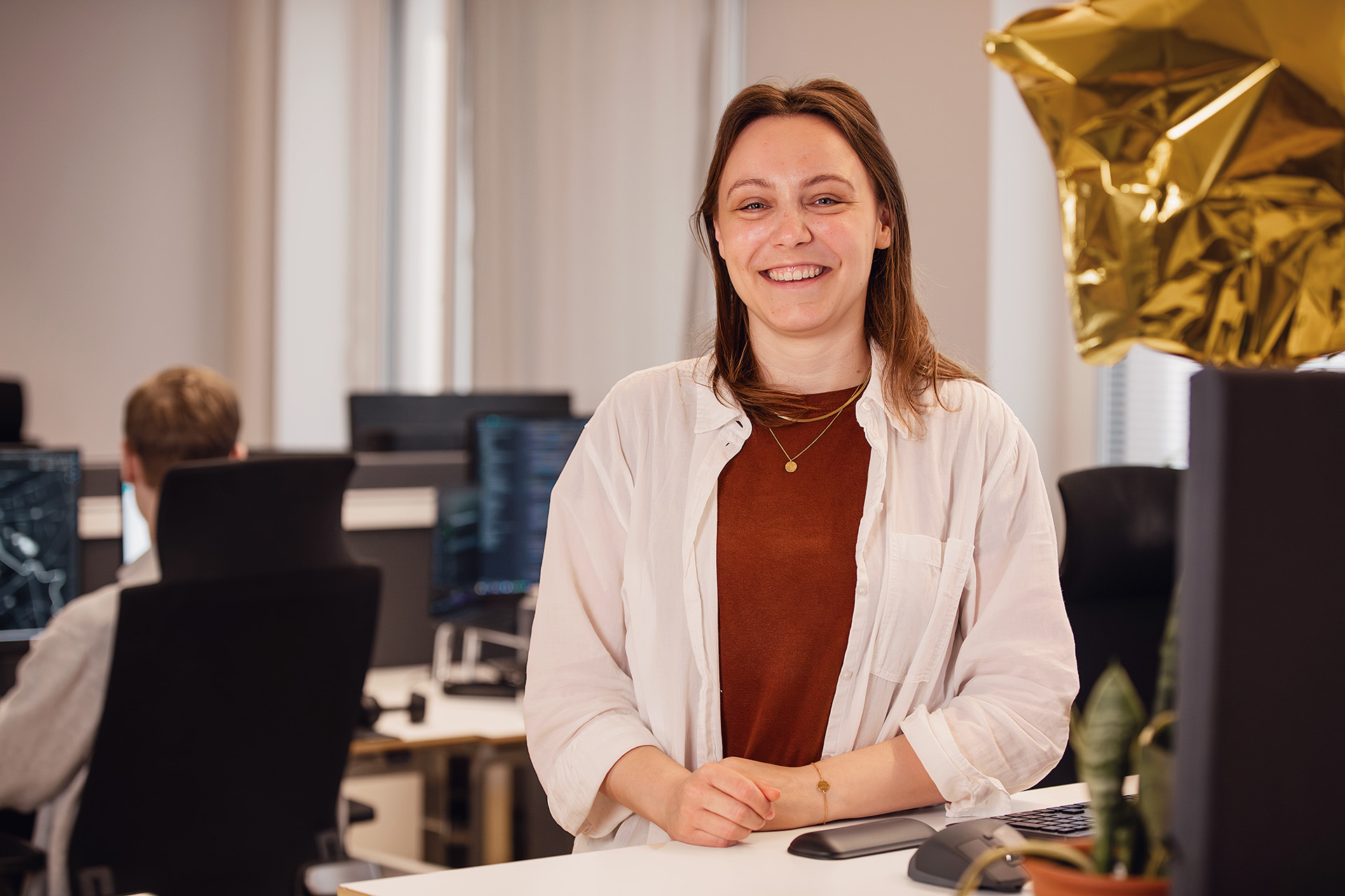 Rebecca står vid ett skrivbord på Gaias kontor och kollar in i kameran och ler. 